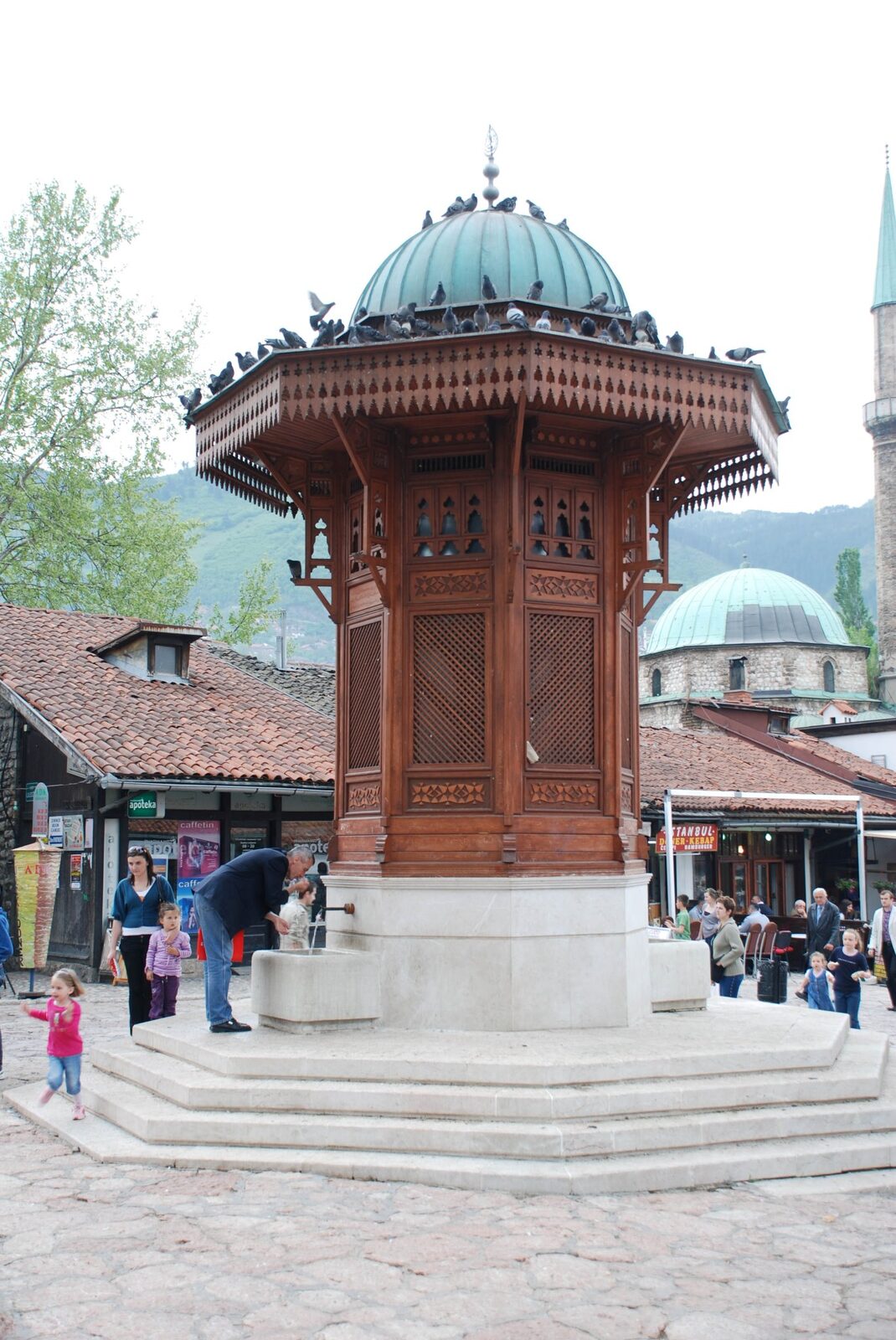 The Sebilj fountain in central Baščaršija, with pigeons descending and tourists feeding them at the stone basin.