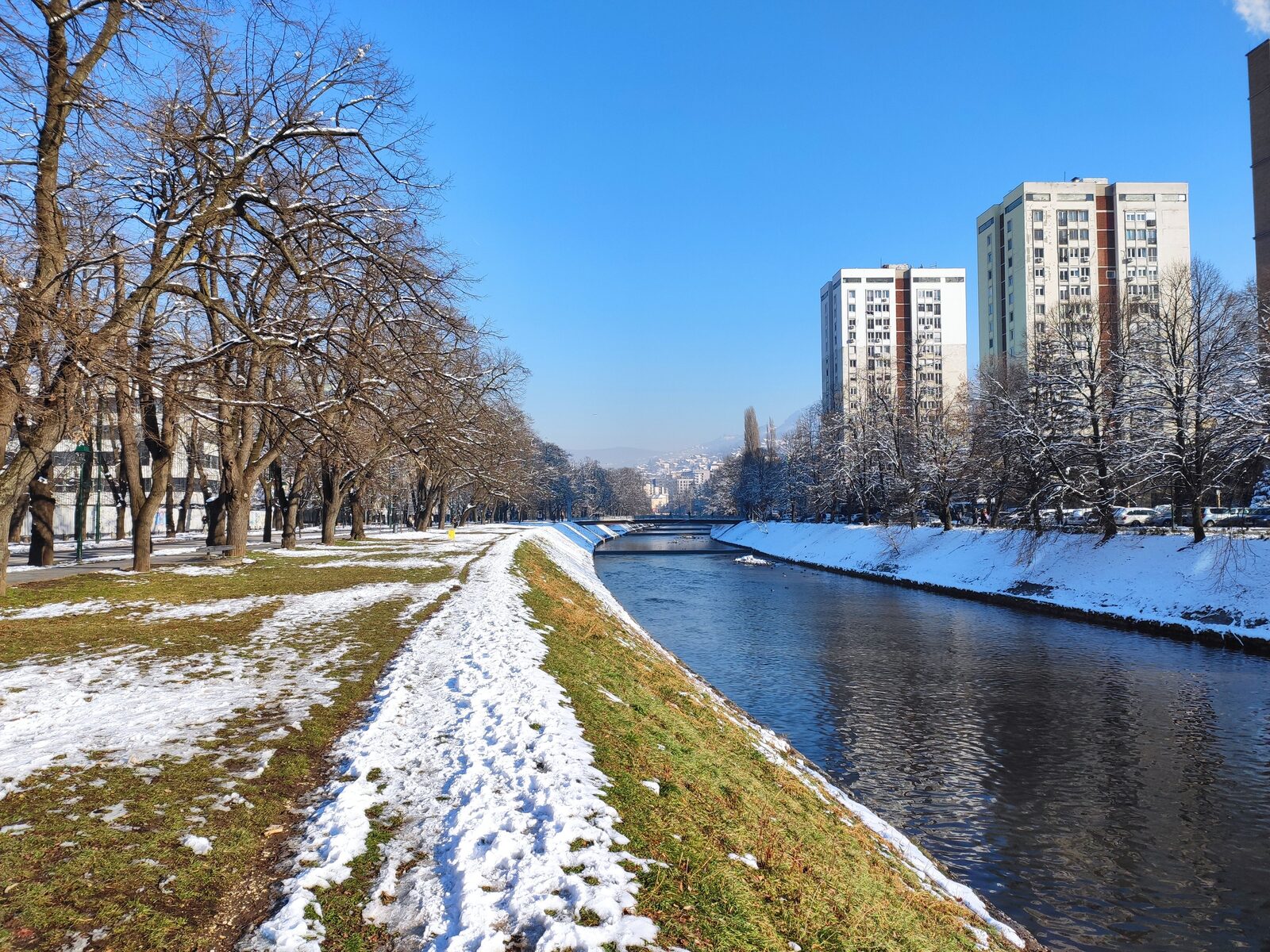 A winter scene along the Miljacka River in Sarajevo, with snow on the embankment walls, bare trees, and the river running dark between the historic buildings. The valley catches snow for most of December through February.