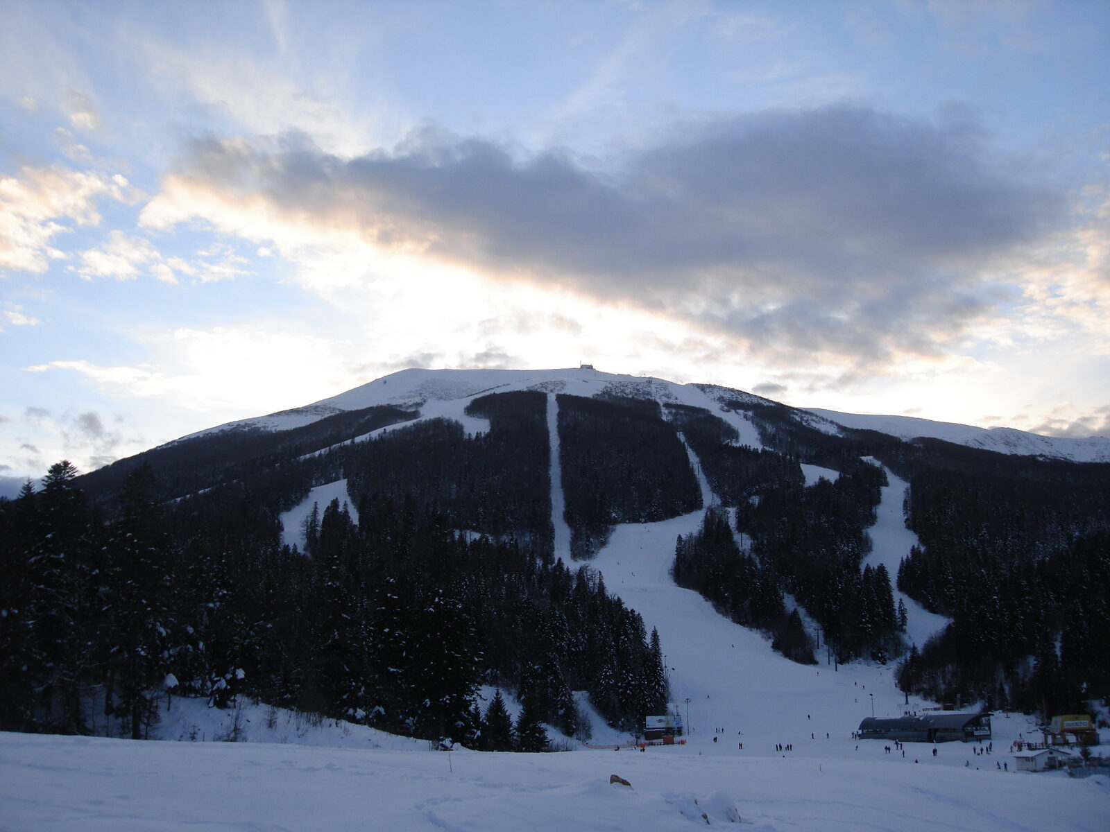 A winter scene on Bjelašnica mountain south of Sarajevo, with snow-covered slopes, pine forest, and the 1984 Olympic ski village in the distance.