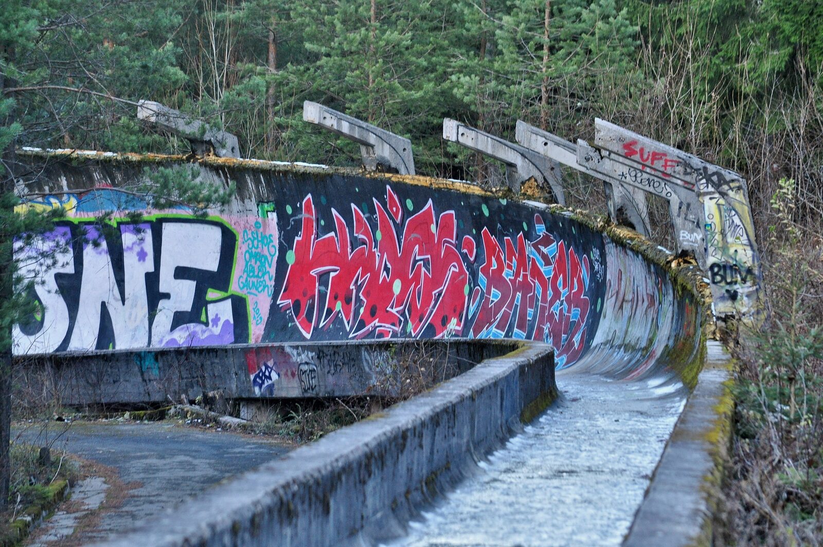 A close view of one of the curves of the abandoned bobsled track on Trebević, with the graffiti-painted concrete chute rising on the outer side, weathered concrete sleepers visible beneath, and pine forest closing in on either side.