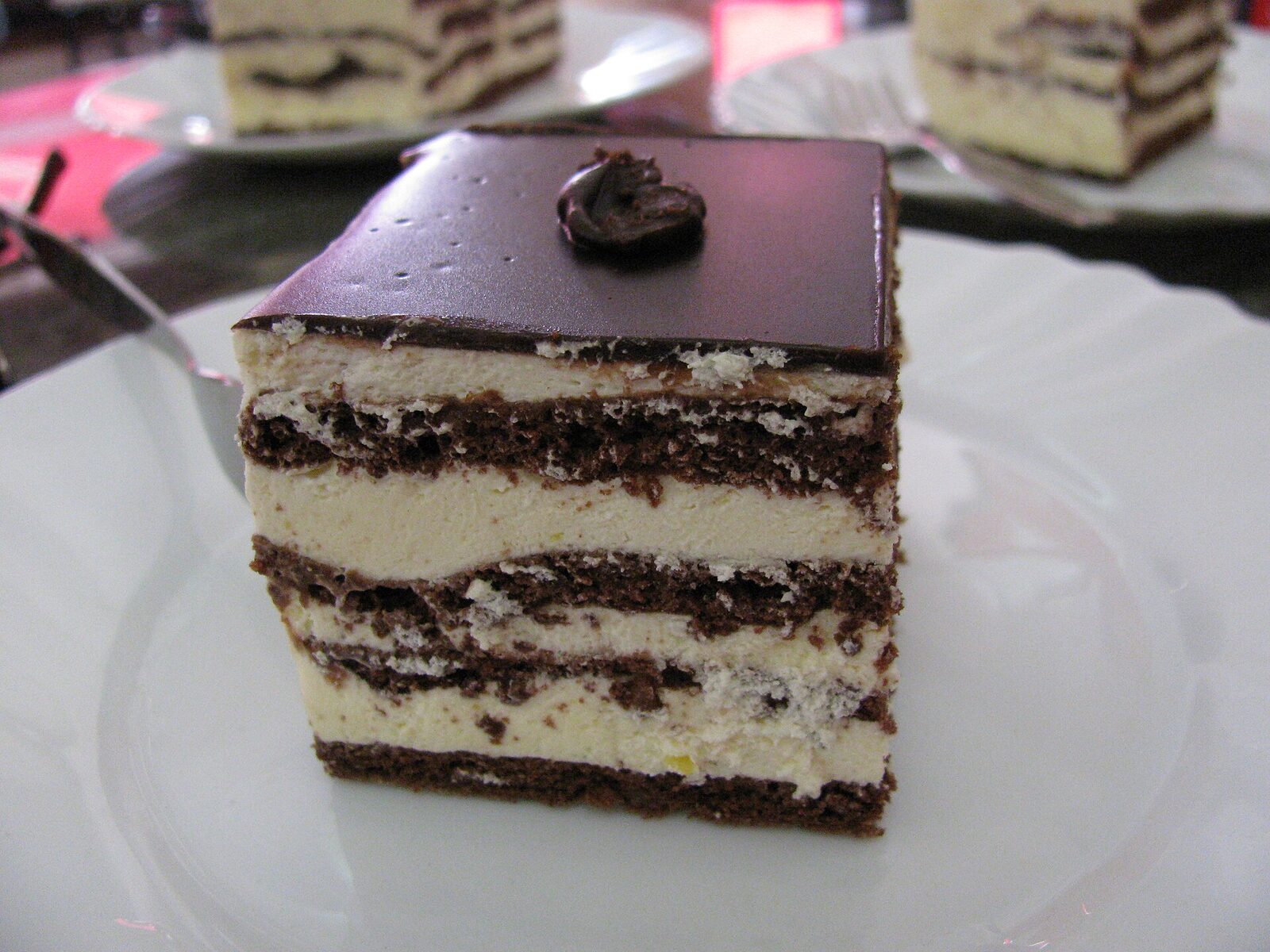 A slice of boem šnita photographed in Sarajevo — a layered cake of chocolate sponge, vanilla cream, and a dark chocolate glaze on top with a small chocolate rosette.