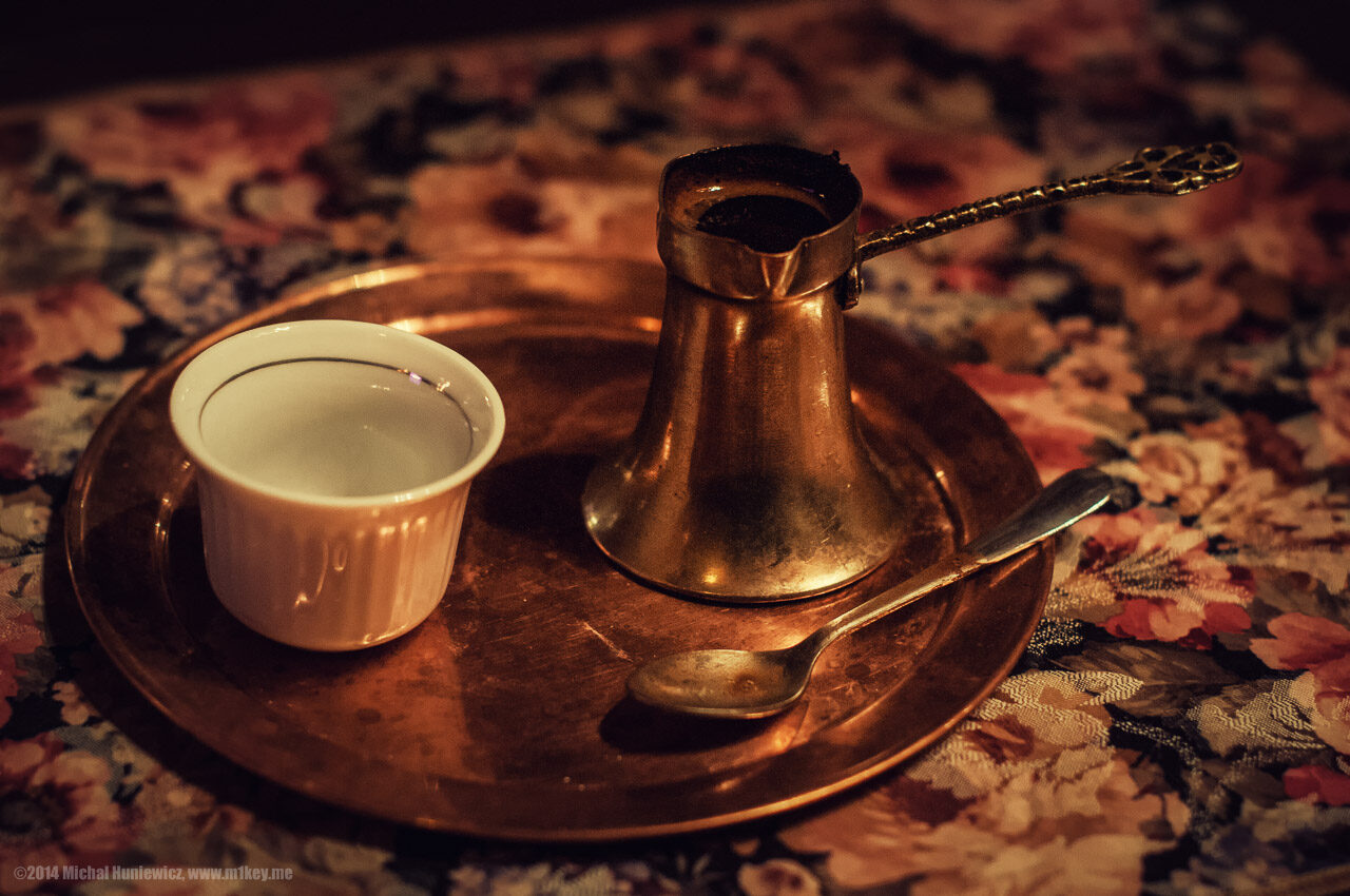 A Bosnian coffee service — a small copper džezva, a fildžan cup, a cube of sugar, and a small glass of water, set on a brass tray on a wooden table.
