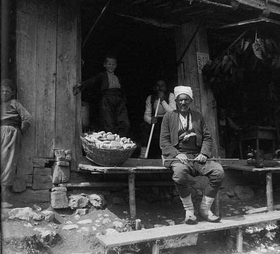 A Bosnian bakery photographed circa 1906 by Rudolf Bruner-Dvořák — a baker in a fez seated on a wooden bench outside his shop, a large woven basket of round somuni beside him, a child in the doorway and an older woman in the shadow of the beam.