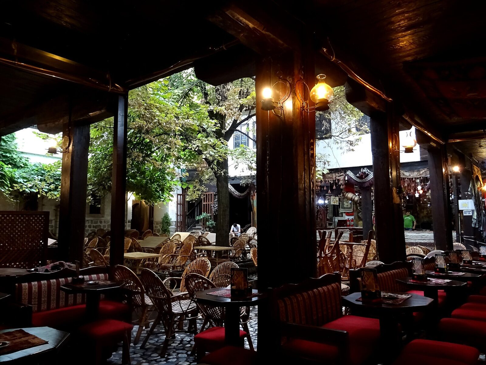 Interior of Caffe Divan at Morića Han in Sarajevo — low red-cushioned Bosnian bench seating along dark timber-post galleries, rattan chairs and small tables in the courtyard beyond, trees visible past the arcade.