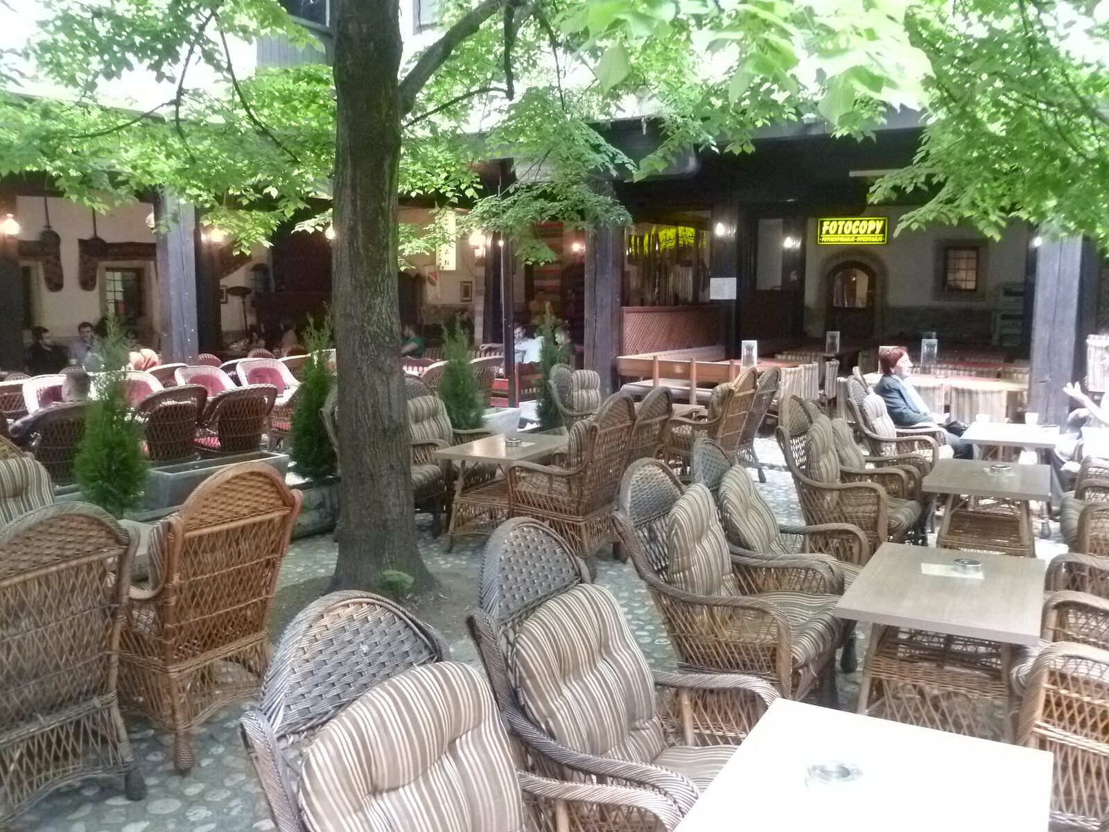 The Morića Han courtyard in Sarajevo — rattan café chairs and small tables under a large tree, cobbled paving, and the dark two-storey Ottoman arcaded gallery visible around the courtyard.