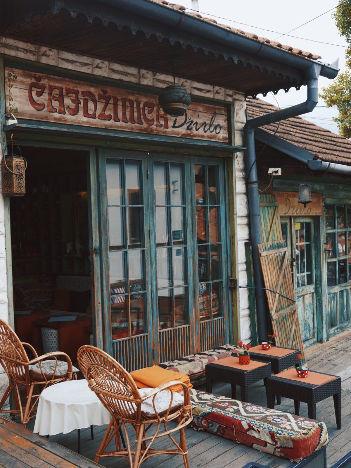 Exterior of Čajdžinica Džirlo — weathered blue-green wooden doors, hand-painted sign, rattan chairs and a kilim-covered bench on a small terrace.
