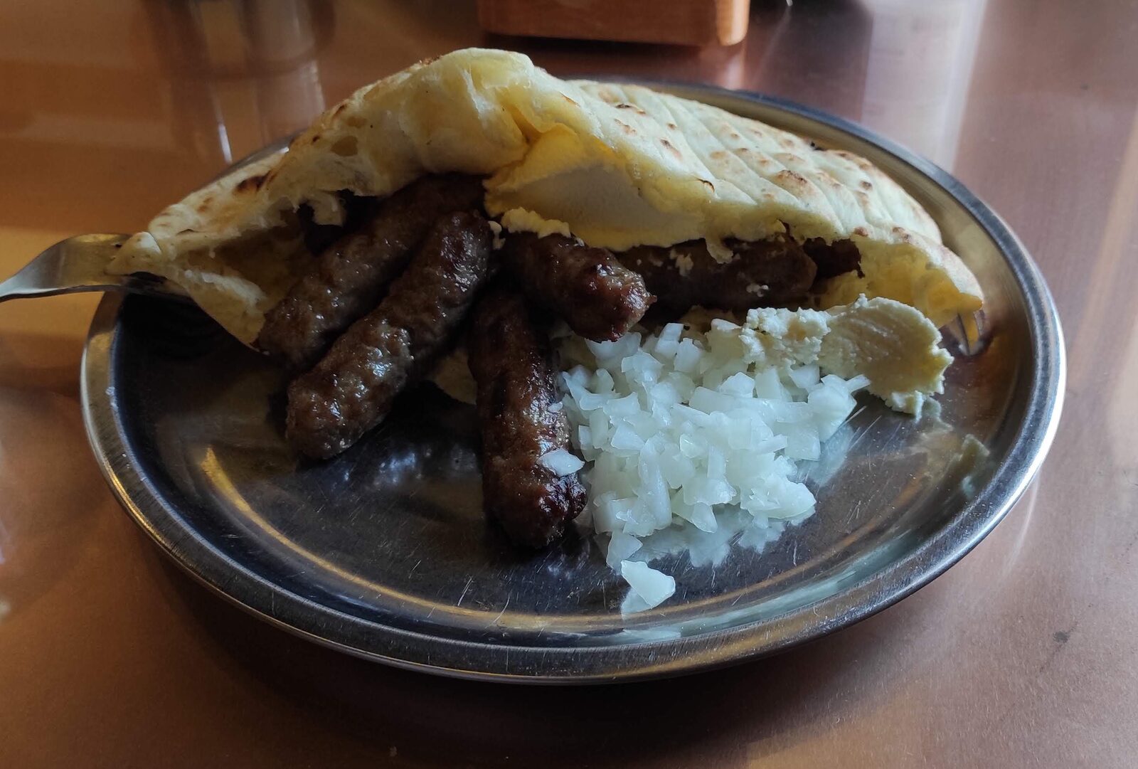 A plate of Sarajevo ćevapi — small grilled minced-meat fingers in a round somun bun, with chopped raw onion and a side of kajmak.