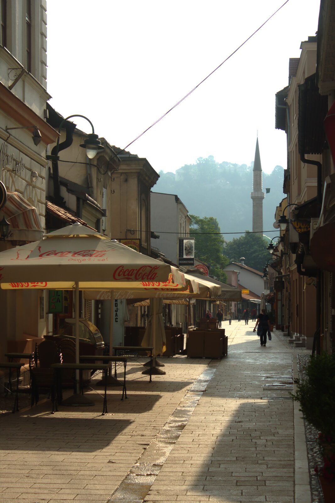A view through Baščaršija toward the Čurčiluk veliki street — a cobbled bazaar lane with low stone shopfronts and pedestrian foot traffic.