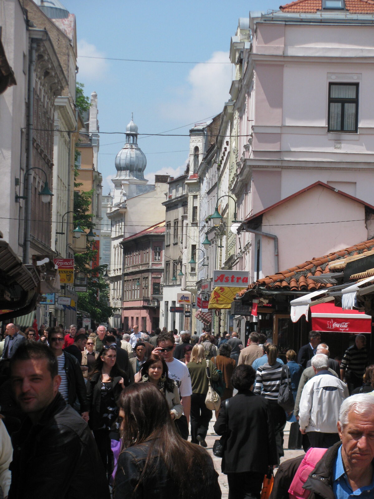 Ferhadija on a busy weekday — pedestrians walking both directions, café terraces full, the Habsburg apartment façades on either side.