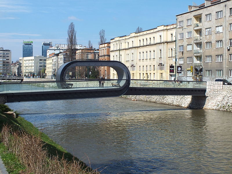 The Festina Lente footbridge over the Miljacka in Sarajevo — a looping steel-and-glass pedestrian bridge next to the Academy of Fine Arts.