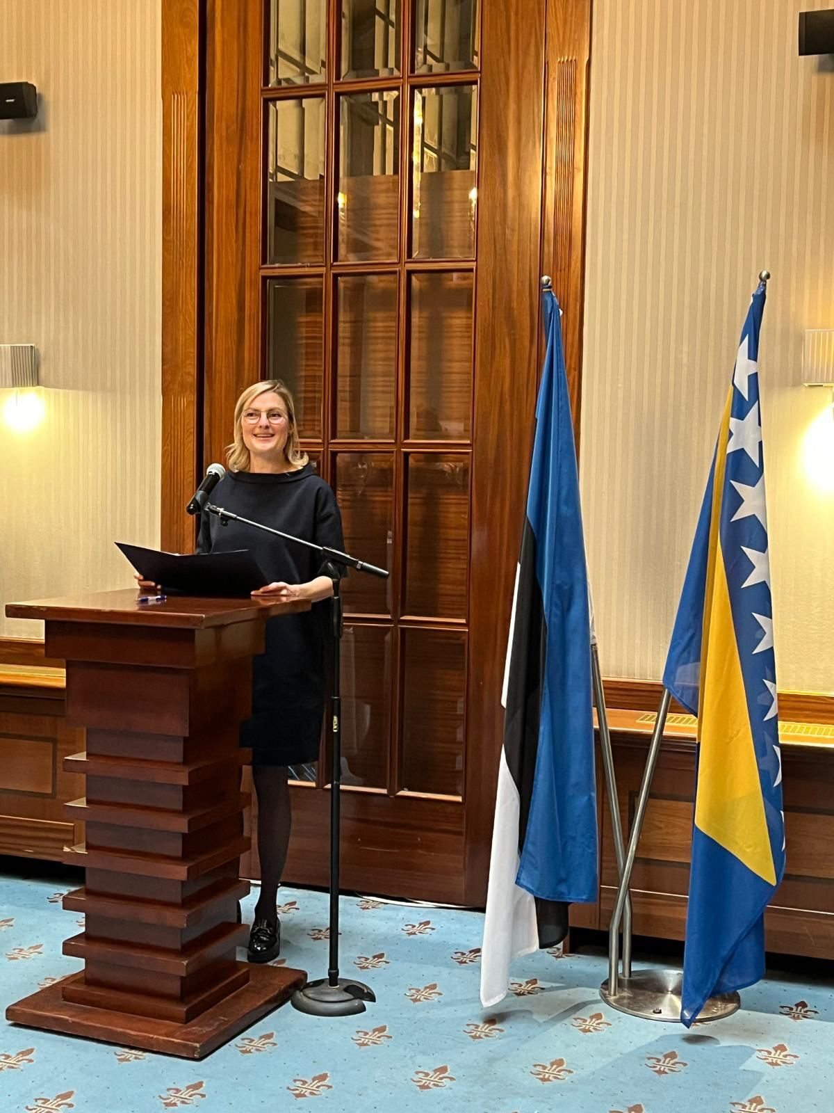 A speaker at a wooden lectern inside Hotel Europe, flanked by the Estonian and Bosnia-Herzegovina flags, in front of a wood-panelled double door and floral-papered walls.