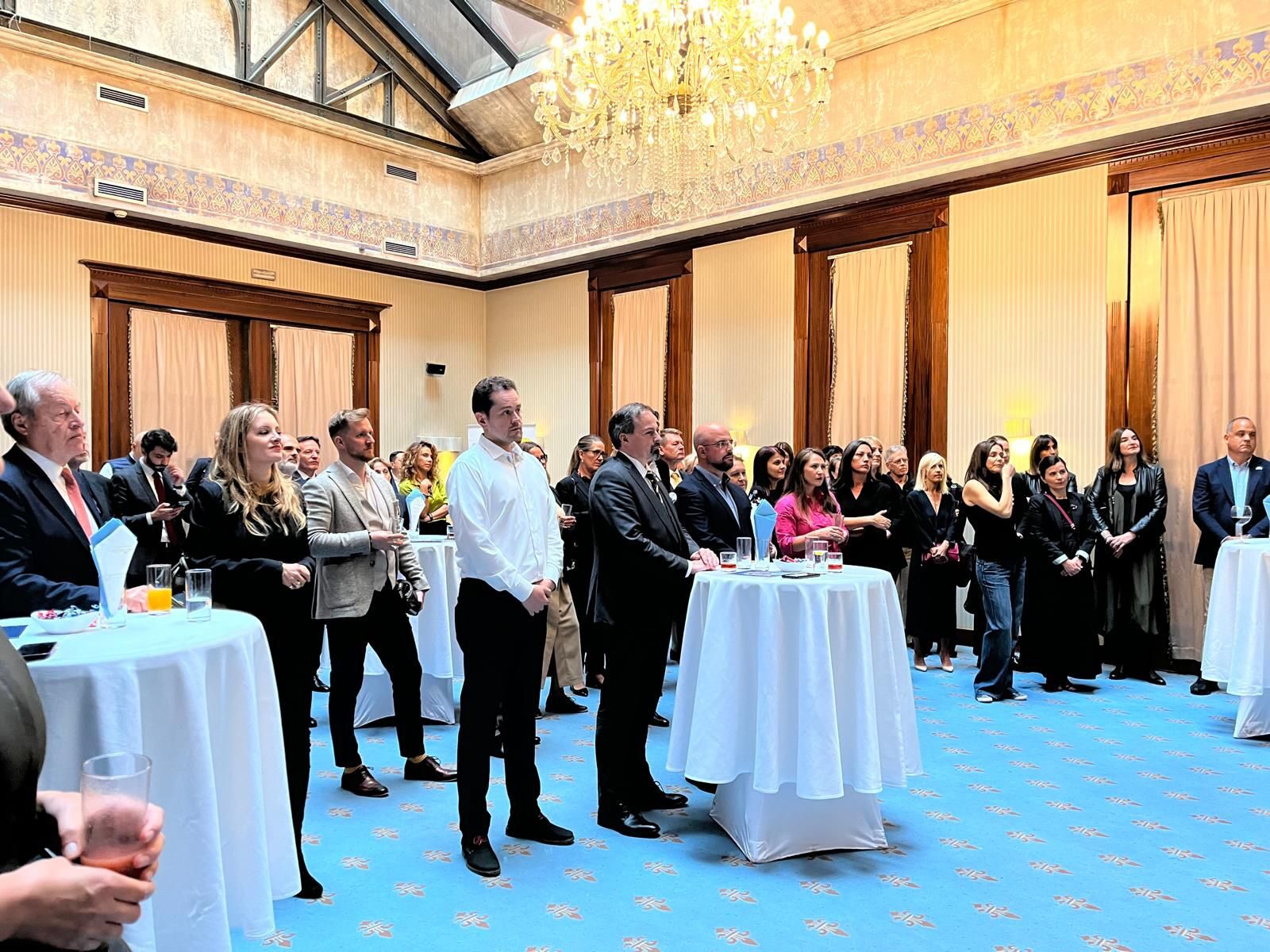 The restored grand event hall of Hotel Europe, with a large crystal chandelier, painted ceiling coffers, tall wood-framed curtained windows, and standing cocktail tables during a reception.