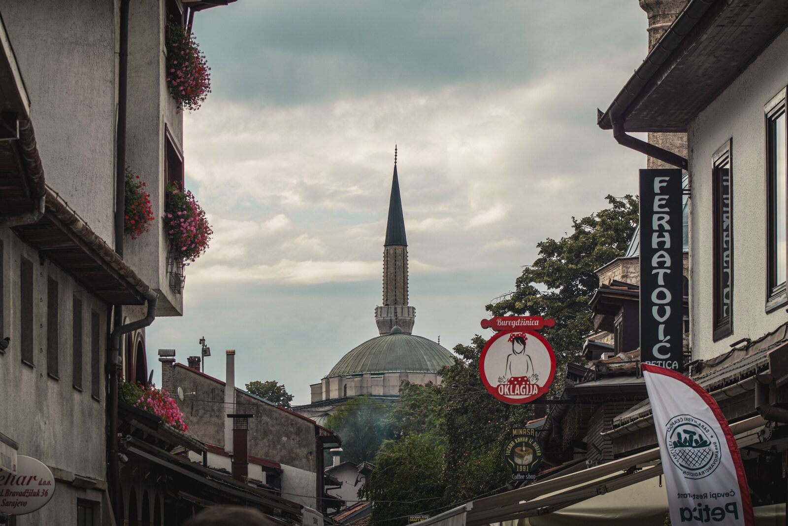 A quiet Sarajevo street at ground level, with cobblestones underfoot and a stone minaret rising above the Ottoman-era buildings in the background. The kind of unhurried moment first-time visitors are surprised to find in a European capital.