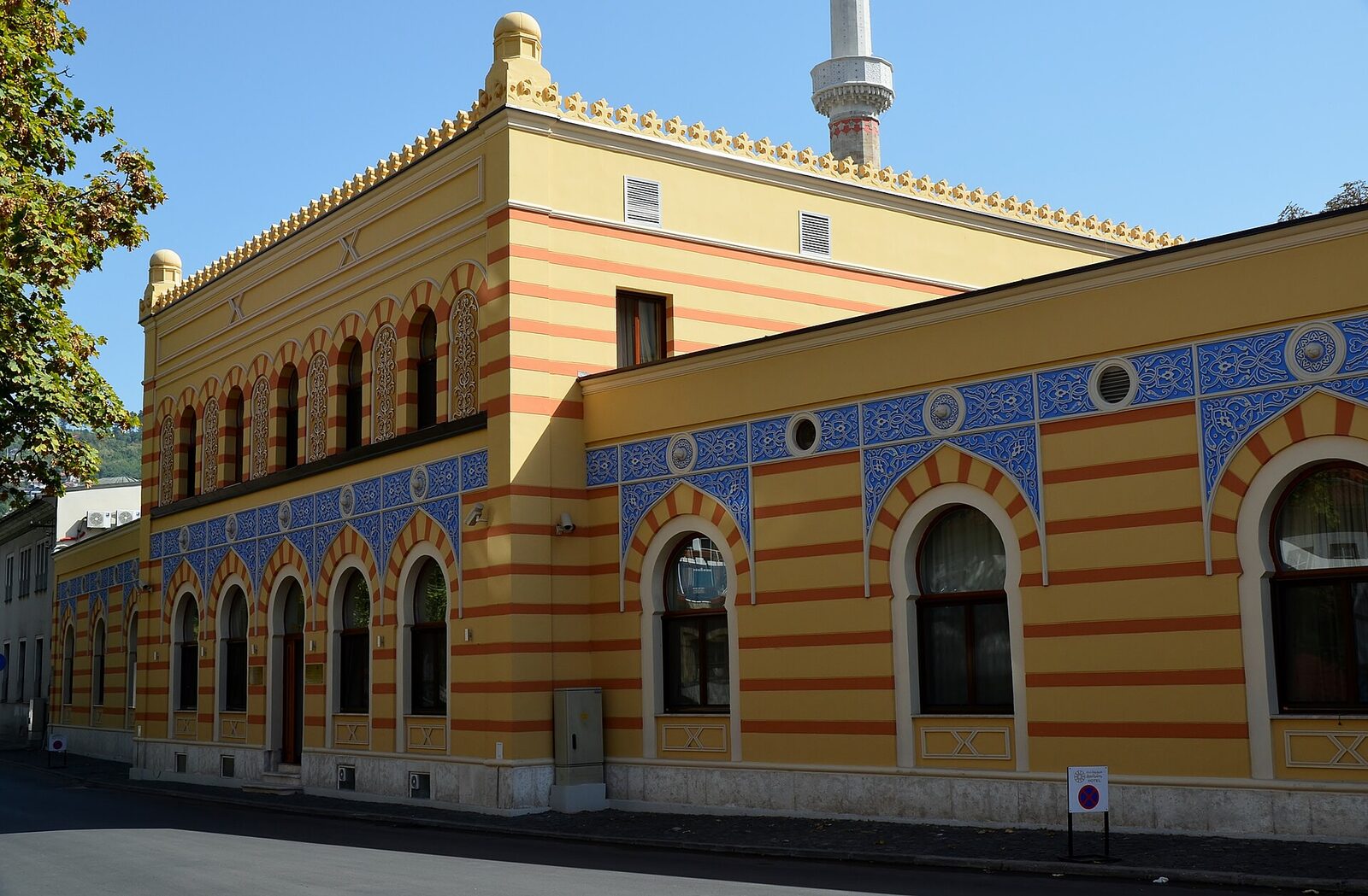 The restored exterior of the Isa-Begov Hamam Hotel in Bistrik, Sarajevo — an ochre-and-blue striped façade with Moorish arched windows and small gold finials along the roofline.