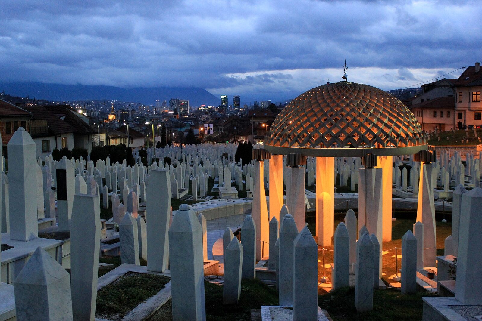 Alija Izetbegović's grave at Kovači Cemetery in Sarajevo — a lattice dome over a stone obelisk, with ranks of white martyr stones around it and the city lights of Sarajevo in the valley behind at dusk.