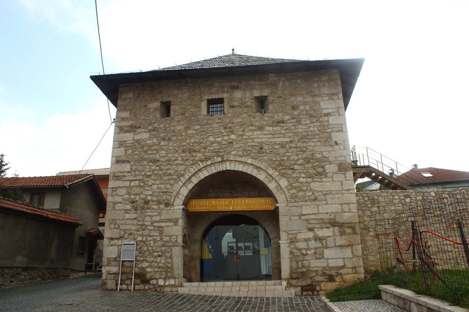 The Alija Izetbegović Museum in Sarajevo, housed in the 18th-century Ploča Gate on the Vratnik defensive walls, with a yellow signboard reading MUZEJ ALIJA IZETBEGOVIĆ 1925 † 2003 in the arched entrance.