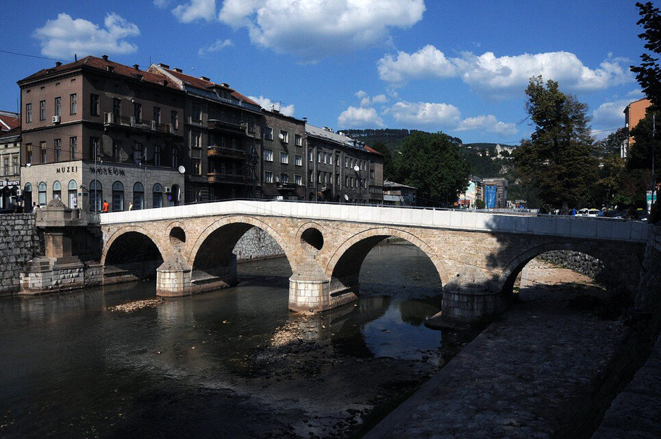 The Latin Bridge over the Miljacka, seen wide from the south bank with the city beyond.
