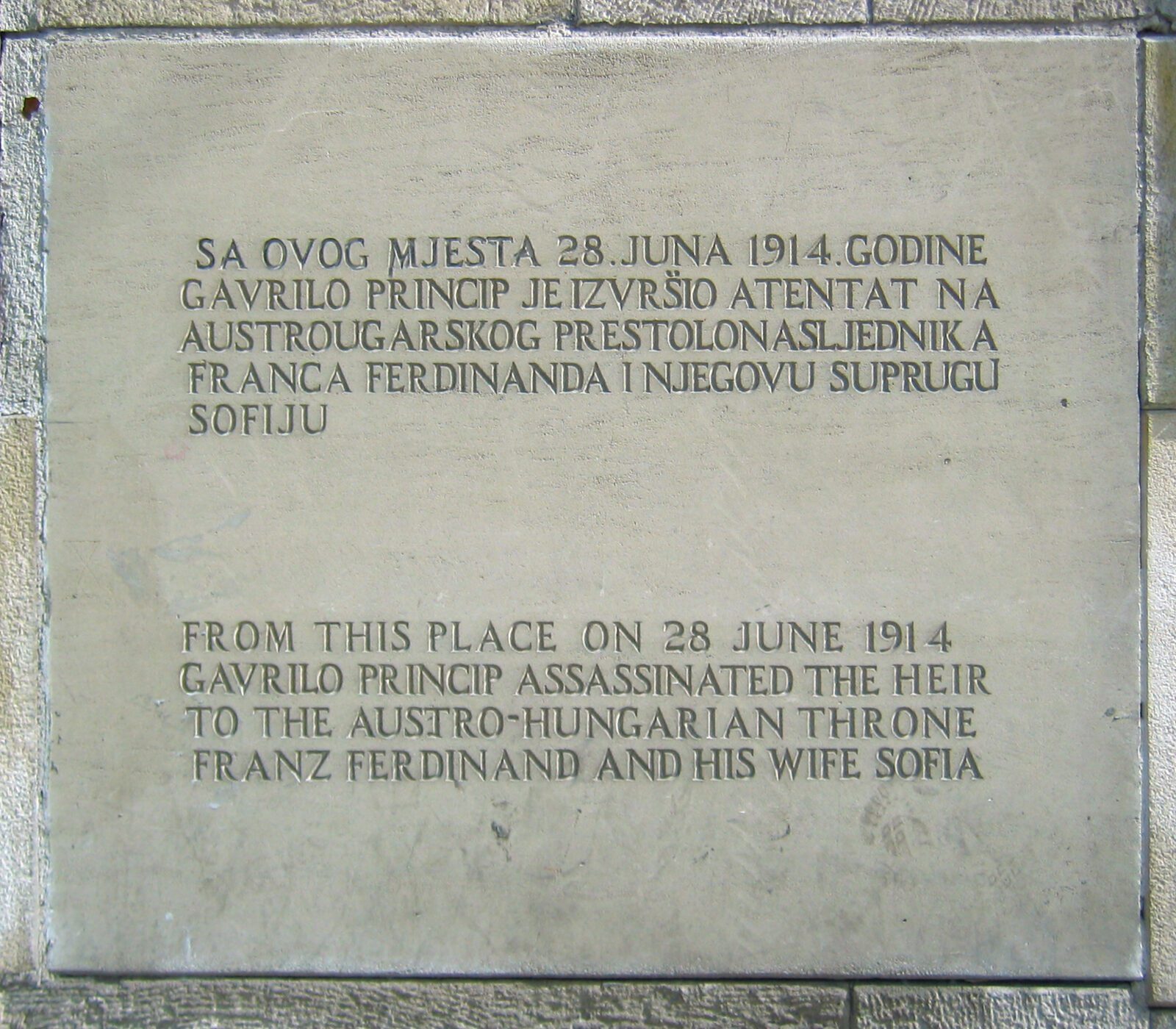 A close view of the stone memorial plaque mounted on the corner wall outside the museum, with an inscription in Bosnian and English commemorating the spot where Gavrilo Princip shot Archduke Franz Ferdinand and Sophie Chotek on 28 June 1914.