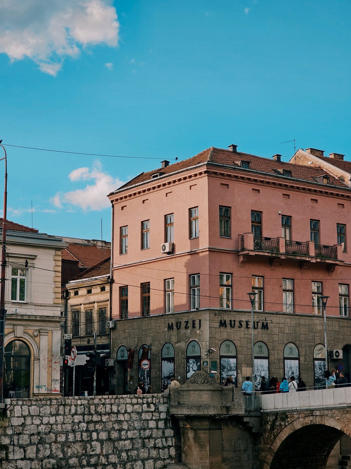 The Museum of Sarajevo 1878–1918 seen from across the Miljacka, with the Latin Bridge in the foreground and the MUZEJ MUSEUM sign visible on the ground-floor facade.