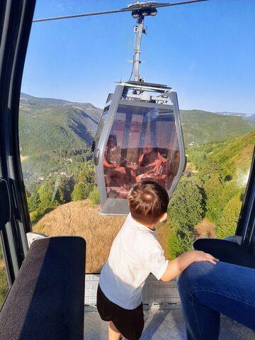 A young child, seen from behind, stands at a cabin window as another cabin passes over green hills.