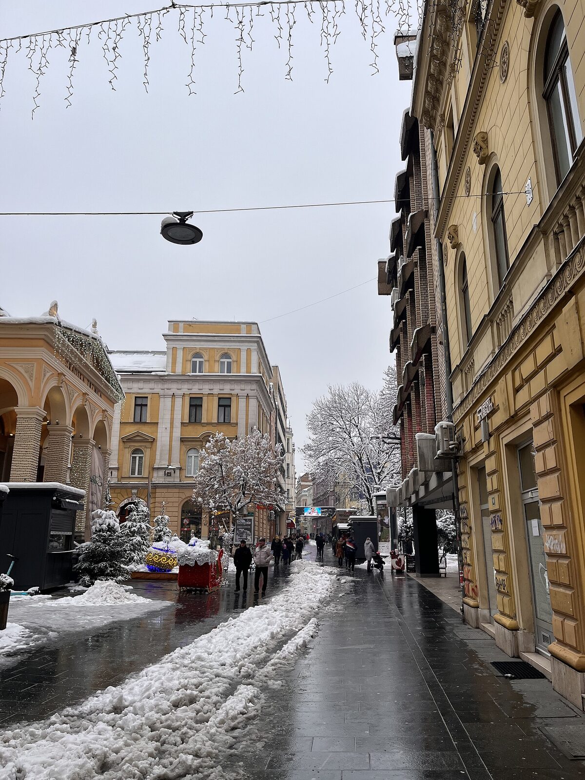 Ferhadija street in central Sarajevo in winter — the pedestrian street the four-faiths walk uses, running west from the Ottoman bazaar past the Meeting of Cultures brass line and on to the Habsburg quarter, with the Sacred Heart Cathedral in the distance.