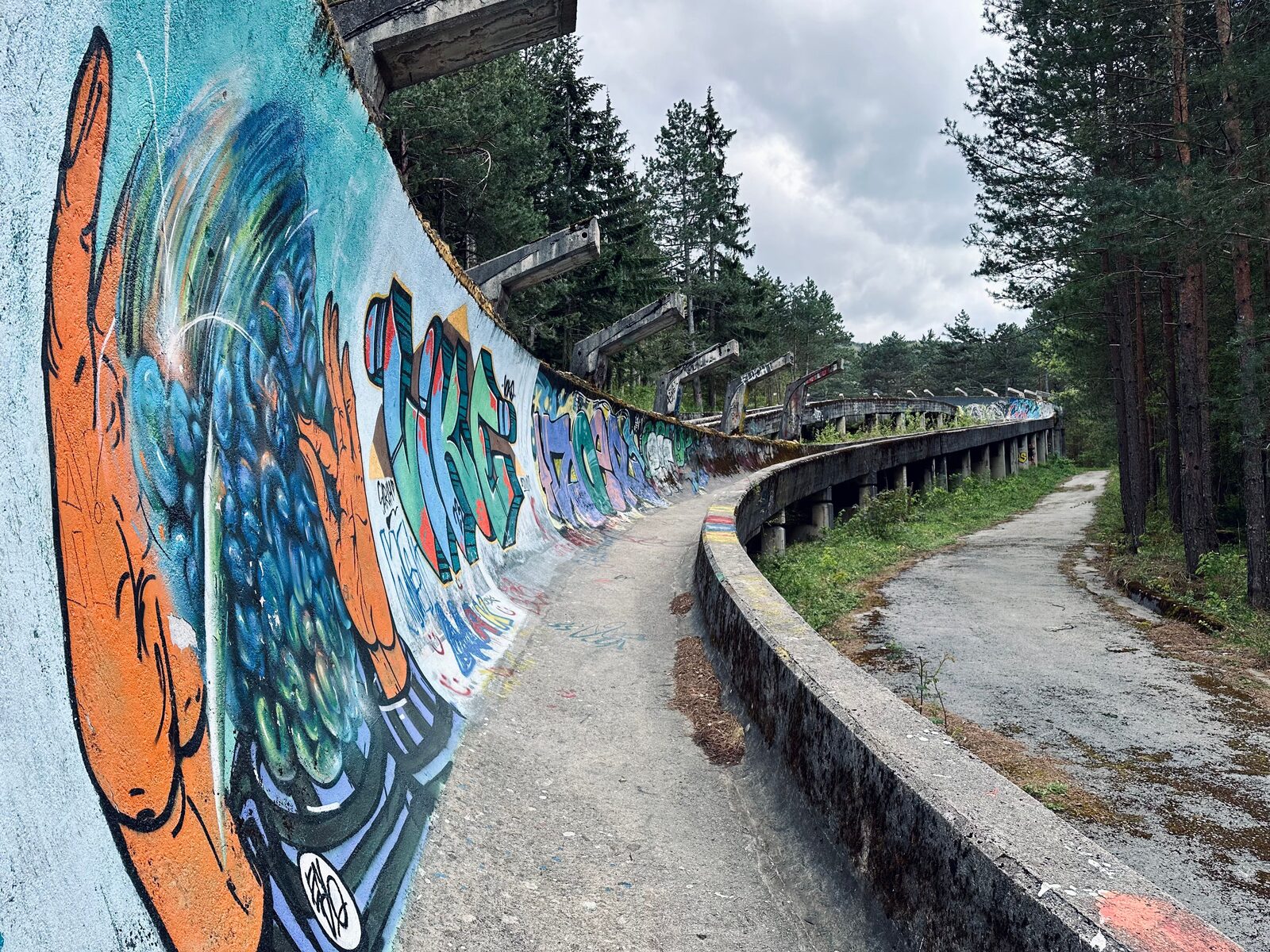 The abandoned 1984 Olympic bobsled track on Trebević above Sarajevo, its concrete walls covered in layers of vivid graffiti art, with lush green vegetation growing through the chute.