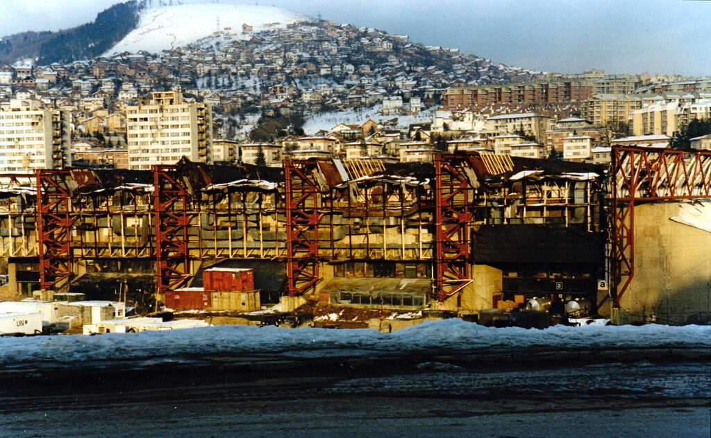 The gutted shell of the Zetra Olympic Hall in Sarajevo during the 1992–1995 siege, the roof collapsed and walls blackened by fire. The hall hosted figure skating and ice hockey at the 1984 Winter Olympics.
