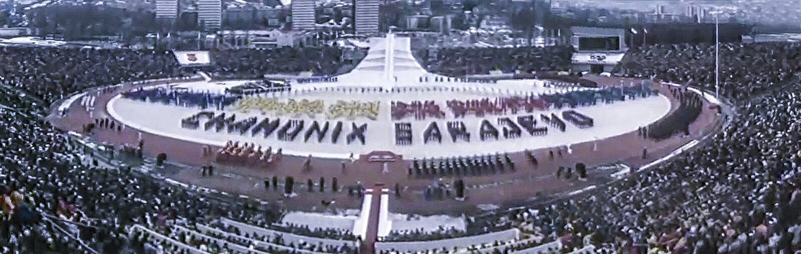 The 1984 Winter Olympics opening ceremony at Koševo Stadium in Sarajevo, with athletes parading on the field and spectators filling the stands. Snow is falling and the Olympic rings are visible above the stadium.