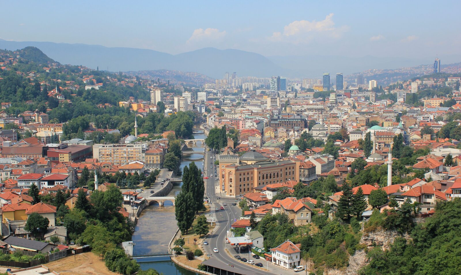 A panoramic view of central Sarajevo, with the Miljacka river, Vijećnica, and the surrounding hills.