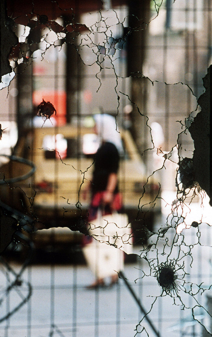 A shattered mirror in a Sarajevo apartment during the 1992–1993 siege — a domestic interior fragmented by shellfire, an image often associated with the city's dark-humour tradition of finding the absurd inside the catastrophe.