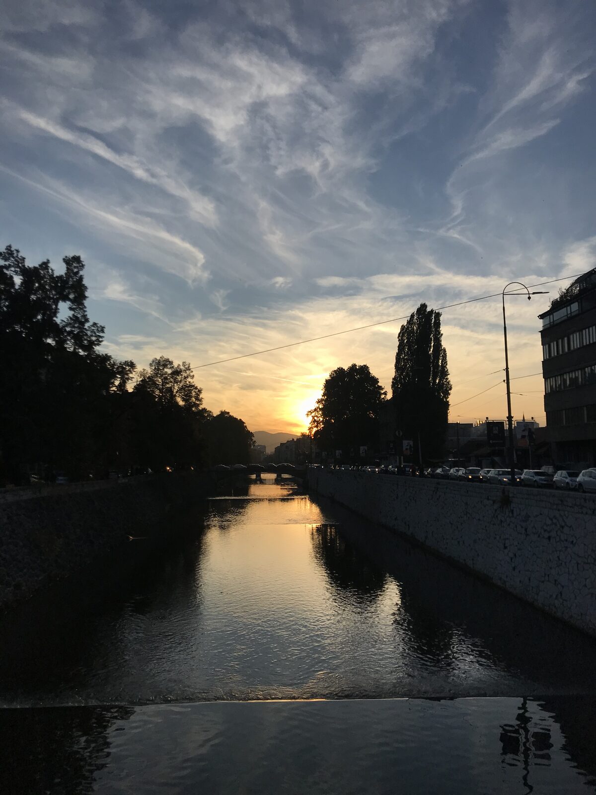 Sunset over the Miljacka river in Sarajevo — the sun dropping behind the western hills, the river reflecting the last light, Lombardy poplars silhouetted against the sky.