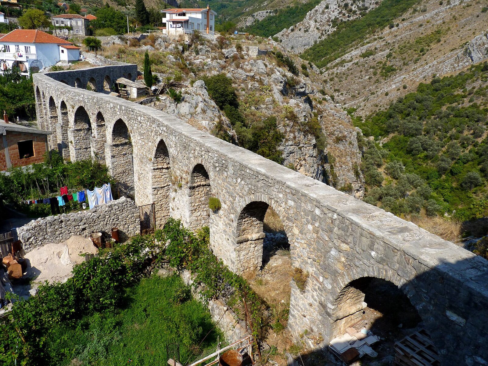 The stone aqueduct of Stari Bar, a row of arches crossing a rocky slope beside houses and gardens.