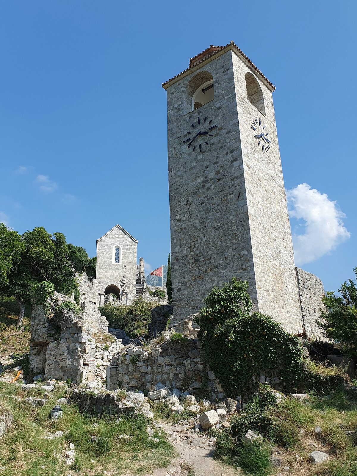 The square stone clock tower of Stari Bar beside a ruined church wall, under a blue sky.