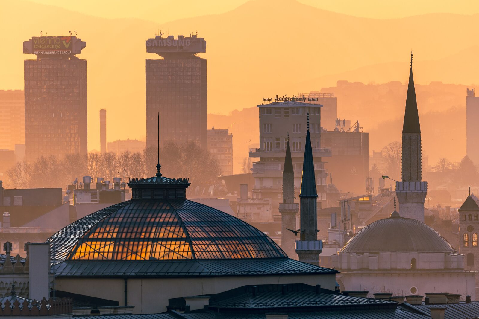 A dusk skyline view of central Sarajevo, with the valley's mosque minarets, tram lines, and Habsburg-era rooftops visible under evening light. The walking tour crosses this corridor east to west along the north bank of the Miljacka.