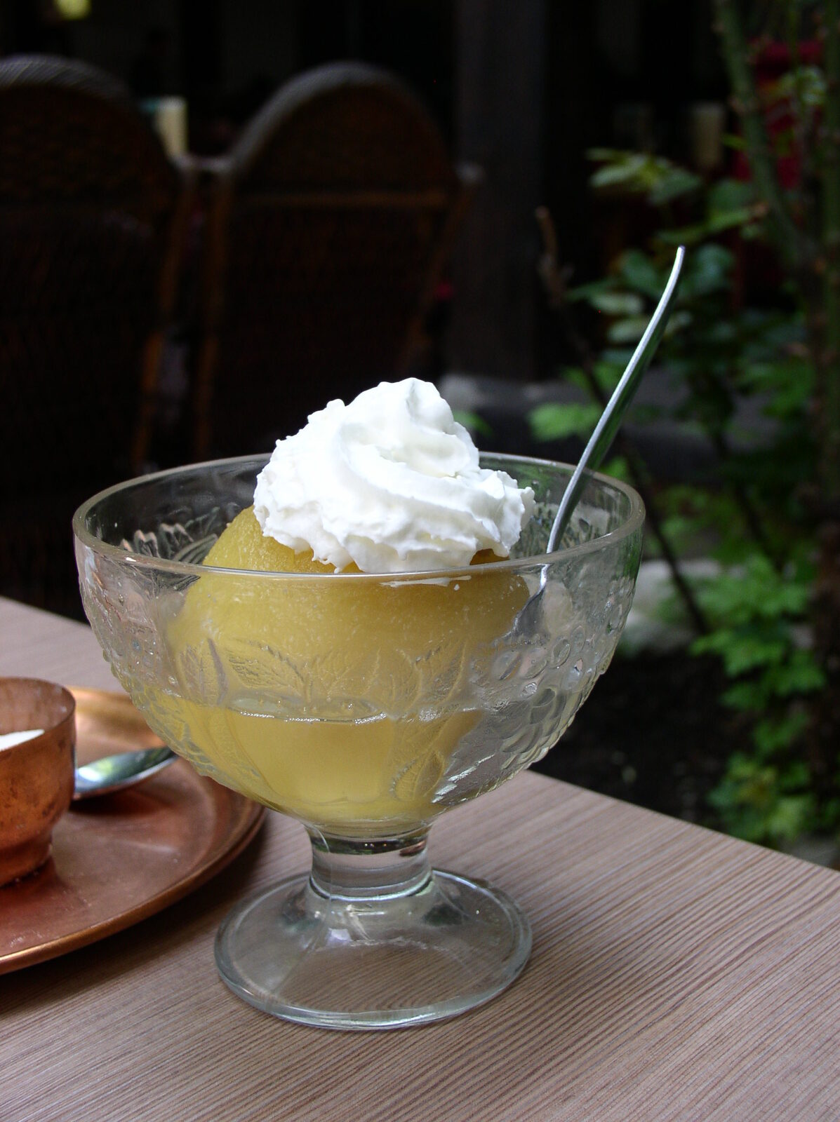 A tufahija served at Morića Han in Sarajevo — a poached whole apple in lemon-sugar syrup, cored and stuffed with a walnut-and-vanilla-sugar filling, topped with a swirl of whipped cream, in a stemmed glass dish beside a copper džezva.