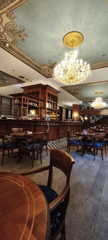 Wooden café tables with navy upholstered chairs under a crystal chandelier and a stencilled, painted ceiling.