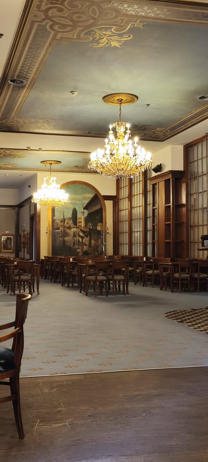 A dining room of wooden chairs under two chandeliers, with an arched mural of an old city street on the far wall.