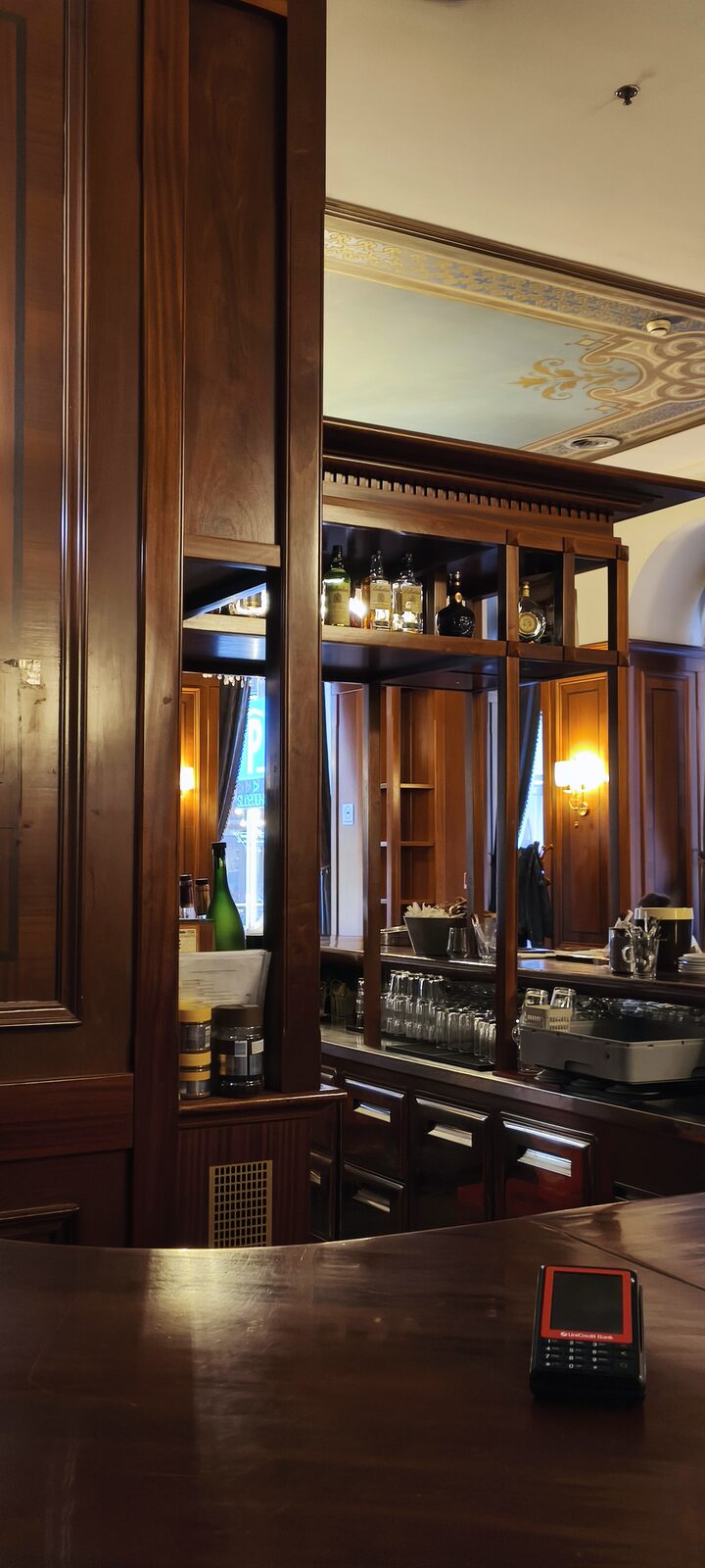 A dark wooden bar counter with shelves of bottles and glasses behind it, under a painted ceiling.