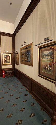 A corridor with striped wallpaper, dark wood panelling and gilt-framed paintings lit by brass picture lamps.