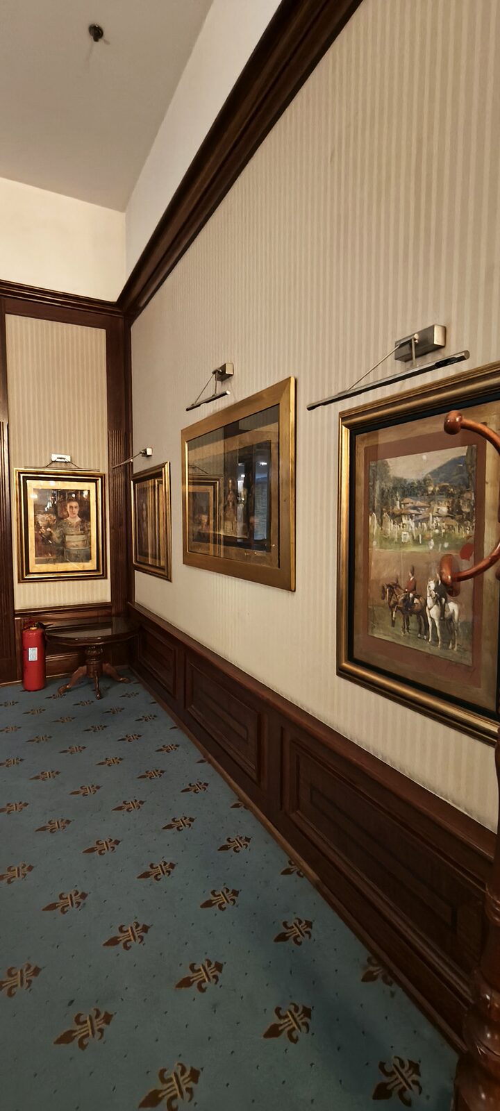A corridor with striped wallpaper, dark wood panelling and gilt-framed paintings lit by brass picture lamps.