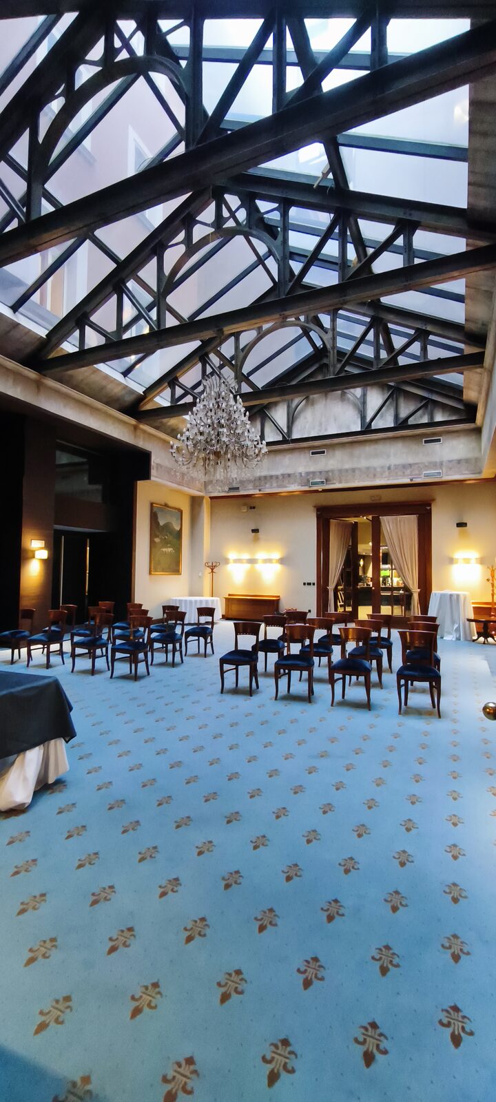 A large hall with a glass roof on steel trusses, a hanging chandelier and rows of dark chairs on blue carpet.