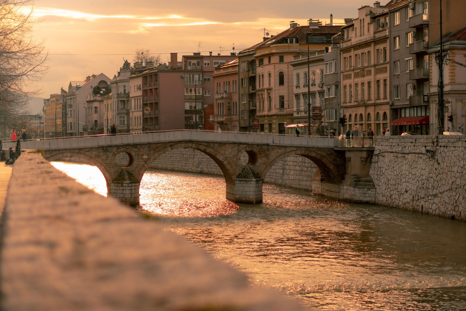 Sunset view of the Latin Bridge over the Miljacka River in Sarajevo, the stone arches catching warm light. The assassination corner is at the far end of the bridge, on the north bank.