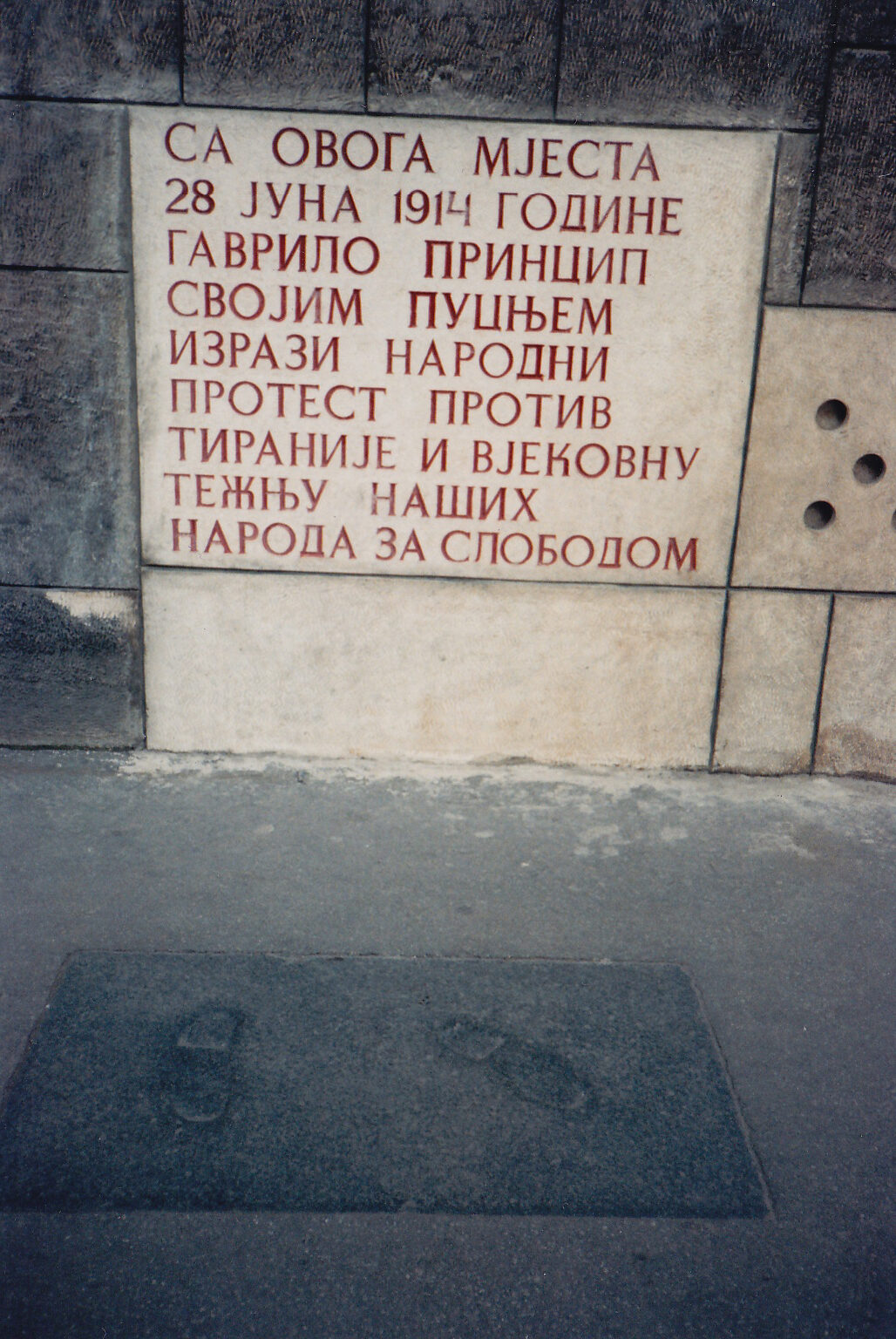 Concrete footprints embedded in the pavement at the corner of the Museum of Sarajevo 1878-1918, next to the Latin Bridge. The footprints mark the spot where Gavrilo Princip stood when he shot Archduke Franz Ferdinand on 28 June 1914. A commemorative plaque is visible on the wall above.