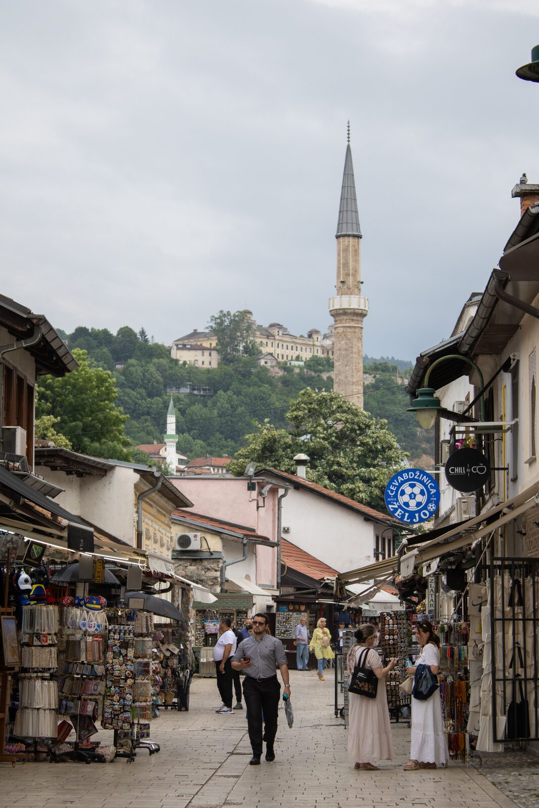 Bravadžiluk street in Baščaršija with the Ćevabdžinica Željo sign visible on the right, a minaret rising behind the low rooftops, and pedestrians walking between the market stalls.